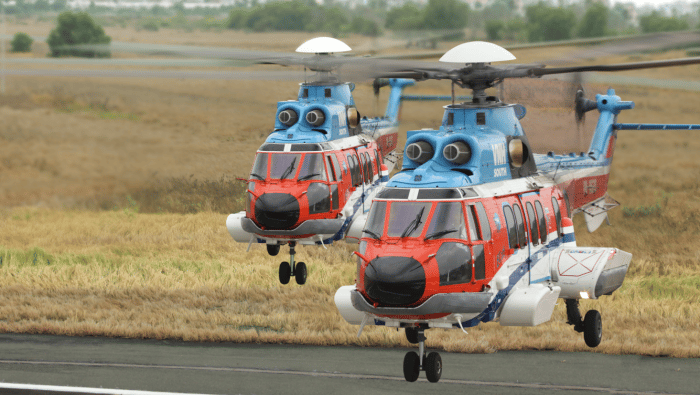 Two VNH-painted orange and blue H225s hovering above tarmac 