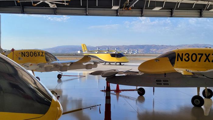 Wisk Aero's fifth-generation Cora prototypes sit in the company's hangar in Hollister, California, as a sixth-generation aircraft crosses the apron outside.