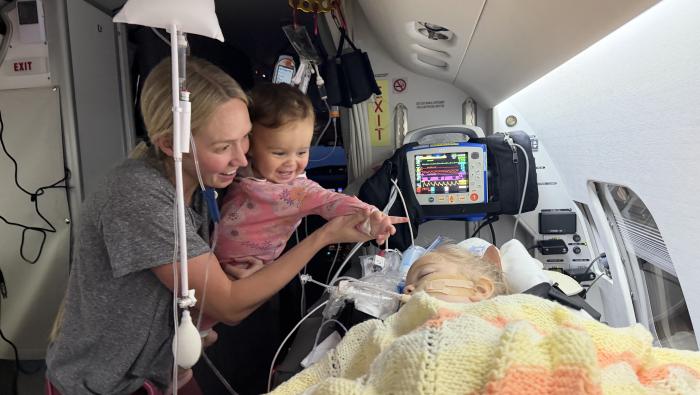 A toddler patient on a bed in a medevac business jet, with a woman and baby visiting with her and smiling