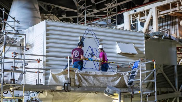 Two technicians on scaffolding paint a blue Artemis logo on a white structure