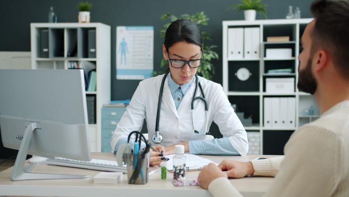 A patient sit's at a doctor's desk while she reviews information on paper in front of her