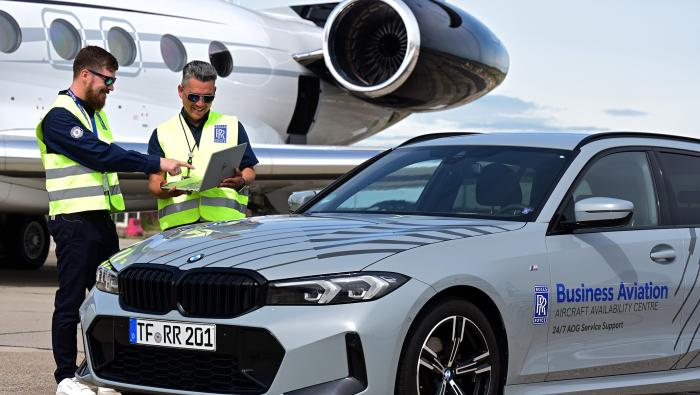 Two Rolls-Royce staff in high-visibility vests look at a laptop beside a business jet and a Rolls-Royce AOG support car.