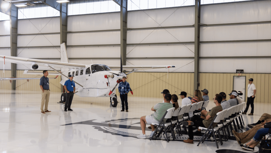 A group of pilots sit in a hangar facing a Kodiak airplane, listening to speakers