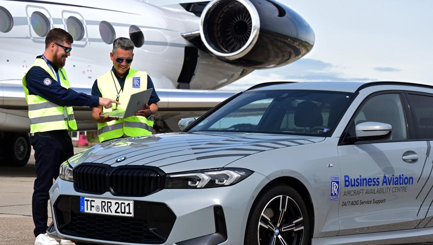 Two Rolls-Royce staff in high-visibility vests look at a laptop beside a business jet and a Rolls-Royce AOG support car.