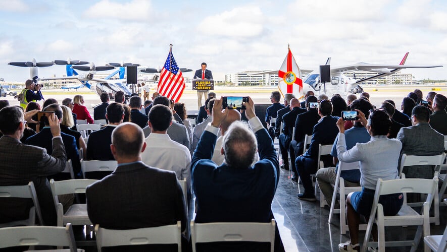 Florida Gov. Ron DeSantis speaks at Tampa International Airport during Florida’s eIPP kickoff event on Oct. 30, 2026, with Joby’s S4 and Beta’s Alia CX300 parked behind the stage.