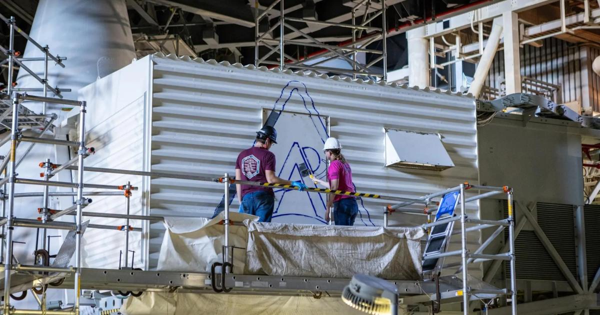 Two technicians on scaffolding paint a blue Artemis logo on a white structure