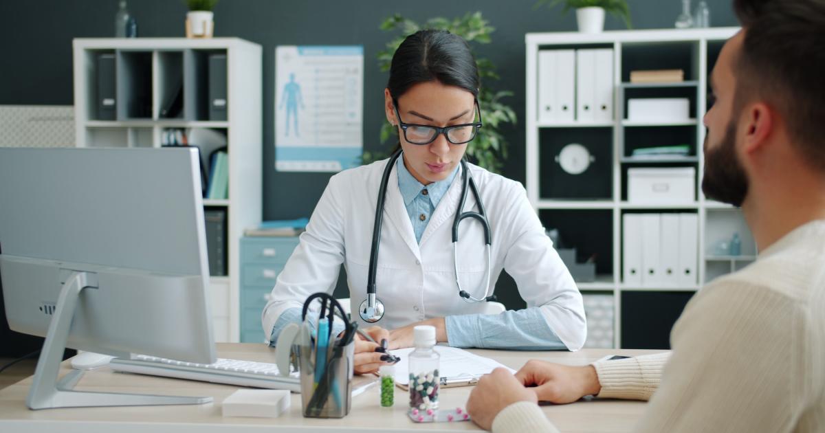 A patient sit's at a doctor's desk while she reviews information on paper in front of her