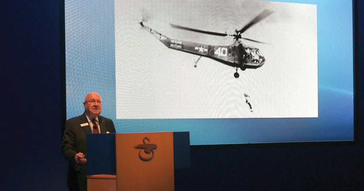 Sergei Sikorsky, behind the podium at Heli-Expo and in the photo behind him, dangling below the helicopter on the rescue hoist during the first flight test of the hoist.